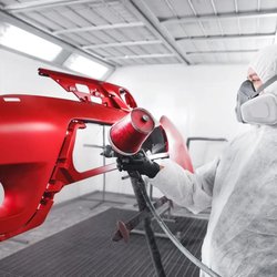 Gervic Auto Body Supply - Auto body technician wearing protective coveralls, gloves, and respirator sprays glossy red paint onto a detached bumper insi...
