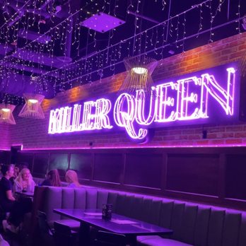 Killer Queen - The Neon Sign on the Front Wall - The image shows the interior of a bar with a prominent neon sign that reads 'Killer Queen' mounted on a brick wall. The ceiling is decorated with string lights and hanging light fixtures, creating a festive atmosphere. There are tables and seating along the wall, with a few groups of people sitting and socializing. The lighting is predominantly purple and pink, enhancing the vibrant ambiance of the venue. - Pizza Near Me - Cincinnati, Ohio