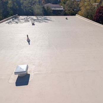 AJ's Professional Contracting - An aerial view of a flat, beige commercial roof featuring multiple vents and a skylight. The roof surface appears smooth and well-maintained. Surrounding trees and a street with parked cars are visible in the background, indicating a suburban or urban setting. - Roofing Near Me