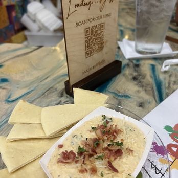 Loading Gastrobrunch - Spinach bacon dip with naan - A plate of spinach bacon dip topped with herbs and bacon bits is placed on a marble table. Next to it are several slices of naan bread. In the background, there is a glass of water with ice, a QR code stand for the menu, and a paper menu. The setting appears to be a casual restaurant or brunch spot with a brick wall visible behind. - Breakfast & Brunch Near Me - St Cloud, Florida