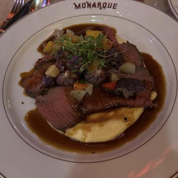Monarque - Steak special - A plated meal at Monarque restaurant includes sliced steak served over a bed of mashed potatoes, topped with a vegetable garnish and microgreens. The dish is presented on a white plate with the restaurant's name, Monarque, printed on the rim. The background shows a table setting with glasses, utensils, and a butter packet. - French Near Me - Baltimore, Maryland