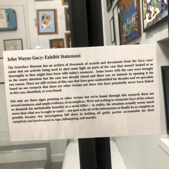 Graveface Chicago - A printed exhibit statement about John Wayne Gacy is displayed on a black stand inside the Graveface Museum, with framed artwork on the wall behind. - Vinyl Records Near Me - Chicago, Illinois