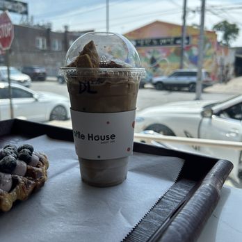 Croffle House - 20 oz. Iced Dalgona Latte - A 20 oz iced dalgona latte in a clear plastic cup with a dome lid and a paper sleeve from Croffle House bakery cafe is positioned on a tray inside the cafe. The tray also holds a pastry topped with blueberries and powdered sugar. The background shows a window with a view of parked cars, street signs, and a mural on a building, indicating the cafe's location in an urban area. - Bakeries Near Me - Flushing, New York