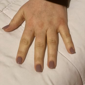 Luxury Nails - The image shows a person's left hand resting on a light-colored fabric surface, possibly a bed or tablecloth. The nails are painted in a taupe or muted brown shade, and the skin appears well-maintained. The person is wearing a dark long-sleeve garment, with the sleeve partially visible at the top of the image. - Nail Salons Near Me - Newberg, Oregon
