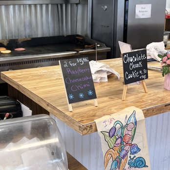Bartels Giant Burger - a kitchen counter with a sign on it - A bakery counter features handwritten signs advertising cookies, including a 'Chocolate Chunk Cookie' priced at $1.50 plus tax and a special offer for '2 for $5.00' on raspberry cheesecake and chimi. A colorful drawing of flowers and a snail is attached to the front of the counter. In the background, a commercial oven or grill is visible, along with a stainless steel refrigerator and some paper napkins or tissues on the counter. - Burgers Near Me - Corning, California