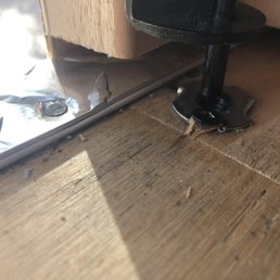 A metal hinge and screw are mounted on a wooden surface, with some debris and a reflective surface nearby.
