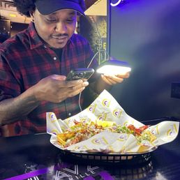 A man in a black Chicago Bulls cap and red plaid shirt is seated at Phlavz Bar & Grille, looking at his phone with a basket of food in front of him.