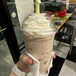 Make your own milkshake! - A person is holding a large cookies and cream milkshake in a clear plastic cup with a dome lid and a yellow straw. The milkshake has visible cookie pieces and is topped with whipped cream. The person is wearing a white watch and has a napkin wrapped around the cup. In the background, there is a wooden table with board games and a window with a view outside.