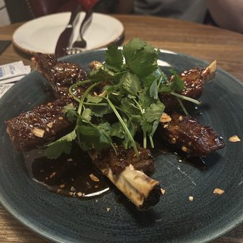 The Dearborn - Sticky Pork Ribs - A plate of glazed pork ribs with a dark, sticky sauce, garnished with fresh cilantro and chopped nuts, is placed on a wooden table in a restaurant setting. In the background, there are empty plates, utensils, and a person taking a photo with a smartphone. The image captures a dining experience featuring a meat dish with Asian-inspired flavors. - New American Near Me - Chicago, Illinois