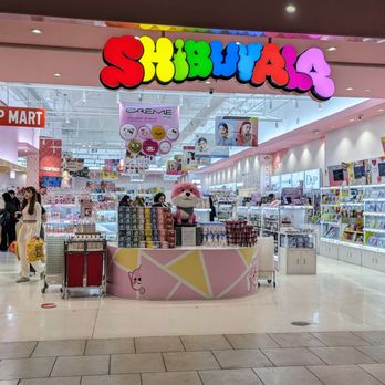 Shibuyala - The image shows the entrance of Shibuya4, a retail store located inside a shopping mall. The store has a colorful, illuminated sign with the name 'SHIBUYA4' in large, multicolored letters. Inside, there is a pink display counter with a plush cat mascot on top, surrounded by various cosmetic and beauty products. The store is well-lit, with shelves and displays filled with items, and several shoppers browsing the merchandise. The mall corridor outside the store is visible, with a few people walking by and carrying shopping bags. - Cosmetics & Beauty Supply Near Me - Arcadia, California