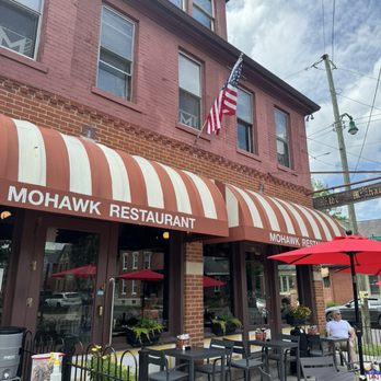 The Old Mohawk - The photo depicts the exterior of Mohawk Restaurant, a multi-story brick building with a red and white striped awning displaying the restaurant's name. Outdoor seating includes tables, chairs, and umbrellas, with a person seated at one table. An American flag is mounted on the building's facade. The scene is set on a sidewalk with a street and parked cars visible in the background, under a partly cloudy sky. - American Near Me - Columbus, Ohio