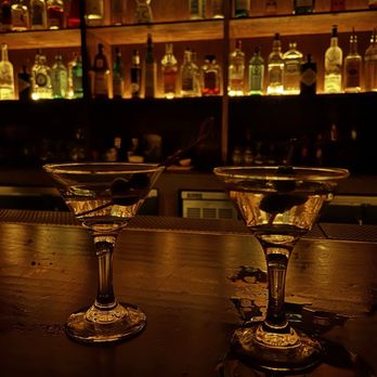 Fox’s Lounge - Martinis - Two martini glasses with olives are placed on a wooden bar counter, with a dark, warm lighting ambiance. The background features shelves filled with various liquor bottles, illuminated from behind, creating a cozy bar atmosphere. The glasses are positioned close together, and the reflections of the glasses and olives are visible on the wet surface of the counter. - Dive Bars Near Me - Miami, Florida