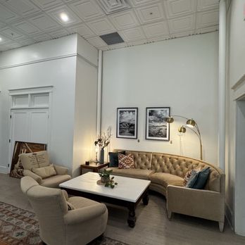 Earl's Cafe - Seating in lobby next to counter for coffee shop - The image shows a living room corner with a curved beige tufted sofa adorned with patterned pillows, a small wooden side table with a lamp and decorative items, and two beige armchairs with pillows. A white coffee table with a small plant is centered in front of the sofa. The room has white walls, two black-and-white landscape photographs on the wall, and a floor lamp with multiple gold-colored shades. The ceiling features decorative square panels with recessed lighting, and the floor is covered with a patterned rug. The overall decor is neutral and stylish, with a cozy ambiance. - Coffee & Tea Near Me - Bisbee, Arizona