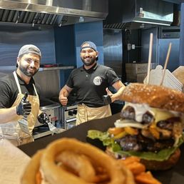 two men standing behind a counter in a restaurant - Two staff members in black shirts and aprons stand behind a food counter, smiling and giving thumbs-up, with a burger and onion rings in the foreground.