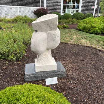 Cape Cod Museum of Art - An abstract stone sculpture is situated in a garden with shrubs and plants, near a building with a window and an 'OPEN' sign. - Museums Near Me - Dennis, Massachusetts