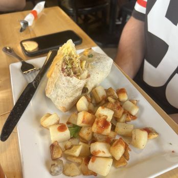 Daily Jam - Breakfast & Brunch Restaurant - The Daily Burrito - A partially sliced burrito filled with ingredients such as meat, cheese, and vegetables is placed on a white square plate. The plate also contains a serving of diced, browned potatoes. The setting appears to be a casual restaurant with wooden tables and black chairs, with a person partially visible wearing a black and white shirt. A smartphone and utensils are also on the table. - New American Near Me - Tempe, Arizona