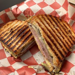 Cubano - A toasted sandwich with grill marks, filled with ham, melted cheese, and pickles, served on checkered paper.