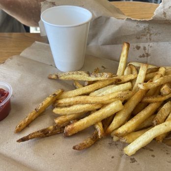 Five Guys - Little Fry - A portion of French fries, some slightly darker and others lighter, are spread on a paper-lined tray. A small container of ketchup is positioned to the left of the fries, and a plain white paper cup is placed behind the fries. The tray is on a wooden table, and part of a person's arm and gray shirt are visible in the background. - Burgers Near Me - Lubbock, Texas