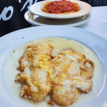 Gino's Restaurant - Filet of Sole Francese - The image features two plates of Italian dishes on a white table. The foreground plate contains two pieces of chicken covered in a creamy white sauce, garnished with small green herbs. The background plate holds a serving of spaghetti with a red tomato sauce. The setting appears to be outdoors, with chairs and a partial view of a building or street in the background. - Pizza Near Me - Brooklyn, New York