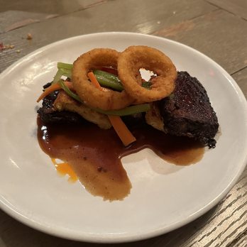 MIX Kitchen & Bar - A white plate holds a serving of fried onion rings placed on top of a piece of grilled meat with a dark sauce. There are also small portions of vegetables, including green and orange strips, garnishing the dish. The plate is set on a wooden table, and the background shows a rustic dining environment. - Cocktail Bars Near Me - Ithaca, New York