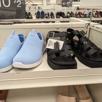 Winners Apparel - Size 11 footwear - A pair of light blue slip-on sneakers with a white sole are placed next to black strappy sandals with a buckle on a retail store shelf. The background features additional shoes arranged on shelves, indicating a footwear section in a retail store. The shelf is white, and the shoes are displayed for sale. - Women's Clothing Near Me - Gloucester, Ontario