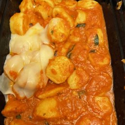 a close up of a dish of food - A black container holds gnocchi coated in a red tomato sauce with visible basil pieces. On one side, there is melted cheese, possibly mozzarella, covering some gnocchi. The dish appears freshly prepared and ready to serve, featuring classic Italian ingredients.