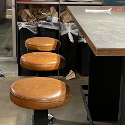 The tables were dirty and the trash cans overflowing - Three wooden bar stools are aligned in front of a counter in a dining area, with trash bins and a paper bag visible in the background.