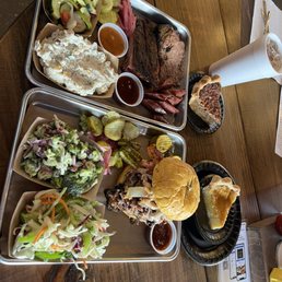 3 Meat - Two metal trays of barbecue food are placed on a wooden table. The top tray contains potato salad, coleslaw, pickles, sliced meats, and sauces. The bottom tray has a salad, pickles, a sandwich with pulled meat and bun, and additional sides. There are also two slices of pie, a large drink in a white cup, and utensils on the table.