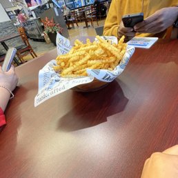 Fried Oreos - A basket of crinkle-cut French fries is placed on a wooden table in a casual restaurant, with people using their phones nearby and a background of large windows and other diners.