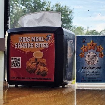 Sharks Burger - A tissue box featuring a Kids Meal Sharks Bites advertisement, including an image of fried chicken bites and a QR code, is positioned on a wooden table. Next to it, a feedback sign with star graphics and a QR code encourages customer reviews. The window behind shows a view of trees and a partly cloudy sky, providing natural light to the scene. - Burgers Near Me - Leander, Texas