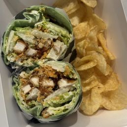 Chicken Caesar Wrap - A chicken Caesar wrap sliced in half, showing lettuce, chicken, and Caesar dressing, served with potato chips in a white takeout box.