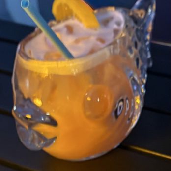 Pesca Vilano - Look at that fish bowl:)) - A fish-shaped glass pitcher holds an orange-colored beverage with ice cubes inside. The pitcher is garnished with a lemon wedge on the rim and has a straw inserted. It is placed on a dark surface, with a dark background behind it. The image is slightly blurry but clearly shows the pitcher and its contents. - Seafood Near Me - St. Augustine, Florida
