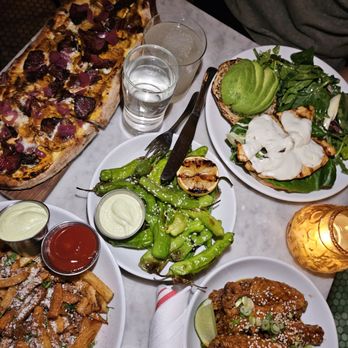 The Binc - A dining table features a pizza with toppings, fried chicken wings with lime, grilled green peppers, and salads, with drinks and a candle. - Cocktail Bars Near Me - Brooklyn, New York
