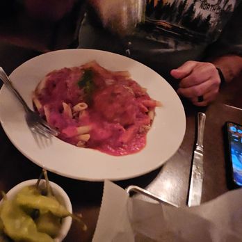 Giacomo's - Meatballs and rigatoni. - The image shows a person wearing a dark shirt with a graphic design, pouring a drink over a plate of pasta with pink sauce and visible toppings. The table has a small bowl of peppers, a fork, a knife, and a smartphone. The setting appears to be a restaurant with dim lighting. - Italian Near Me - Melrose, Massachusetts