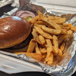 A takeout meal features a burger with a glossy, toasted bun and a beef patty, topped with sliced onions and pickles. Next to the burger, there is a portion of French fries seasoned with black pepper, served in a container lined with aluminum foil. The meal is presented in a foam container, likely for quick service or delivery.