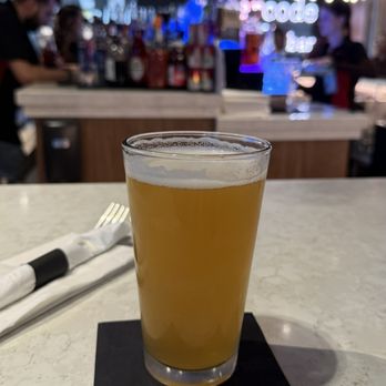 Beercode Kitchen & Bar - Sam Adams - A clear glass of beer with a light golden color and a thin foam head is positioned on a black napkin on a white marble table. The background features a bar area with various bottles, bartenders, and patrons, with dim lighting and a neon sign. The setting appears to be a modern bar or restaurant. - Breakfast & Brunch Near Me - Windsor Locks, Connecticut