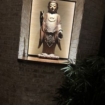 Red Pine - A wooden Buddha statue is positioned in a niche with brick walls, illuminated by a spotlight from above. The statue depicts Buddha standing with one hand raised and the other lowered, on a small platform. The setting appears to be indoors, with a plant partially visible in the lower right corner. - Chinese Near Me - Boca Raton, Florida