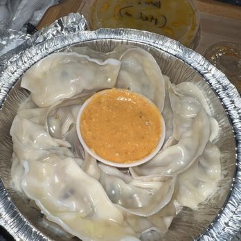 The Punjab - 9 Piece Chicken Momo - The image shows a round aluminum tray filled with steamed chicken momos, arranged in a circular pattern. A small cup of orange-colored dipping sauce is placed in the middle of the momos. In the background, there are additional containers with sauces or broths, and the setting appears to be a food service area, likely at The Punjab restaurant in Johnston, RI. - Indian Near Me - Johnston, Rhode Island