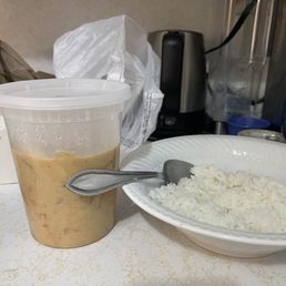 30. Yellow Curry - A plastic cup containing Thai iced tea with a lid and a bowl of cooked white rice with a metal spoon are placed on a table. The background includes a plastic bag, a black appliance, and a blender, indicating a casual dining or kitchen environment.
