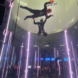The image shows an individual in a skydiving suit and helmet performing indoor skydiving inside a glass tunnel. The person appears to be in free-fall, with their body positioned horizontally and arms extended. The tunnel is illuminated with vertical purple lights, and a group of people can be seen observing through the glass at the bottom. The facility's logo, 'iFLY,' is visible on the ceiling of the tunnel.