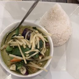 Green Curry - A dish featuring vegetable curry with various vegetables such as eggplant, bell peppers, and carrots, topped with shredded cheese. The curry is served in a small white bowl placed on a square white plate, alongside a cone-shaped mound of steamed white rice. A metal spoon rests in the curry bowl, and the setting appears to be on a wooden table.