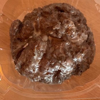 Sweet Circle - Apple Fritter - A chocolate glazed donut with a shiny chocolate coating and white icing drizzle on top is shown inside a clear plastic container. The donut appears to be large and has a textured surface with some uneven areas. It is placed on a wooden surface, and the container is open at the top, revealing the donut clearly. - Donuts Near Me - Schererville, Indiana