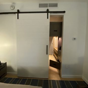 Canopy by Hilton Ithaca Downtown - A white sliding barn door with black hardware is partially open, revealing a hallway or room with wooden flooring, a piece of furniture, and a patterned wall. The door is mounted on a black rail system, and the surrounding wall is painted white. The room appears to be part of a modern interior space. - Hotels Near Me - Ithaca, New York