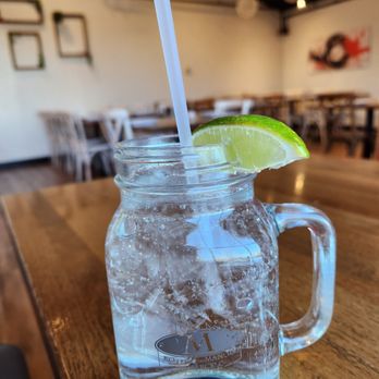 Boteco Do Manolo - A glass mason jar with a handle contains sparkling water, garnished with a lime wedge and a straw, on a wooden table in a restaurant. - Brazilian Near Me - Framingham, Massachusetts