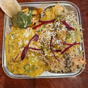 Shimla Peppers - Kadi Pakora & Rice - A metal tray holds a variety of vegetarian Indian dishes, including rice, curry, and vegetable preparations, garnished with shredded cheese and red chili peppers. A papad is placed on the side of the tray, and the tray is set on a wooden table. The dishes appear to be part of a vegetarian meal served at an Indian restaurant. - Indian Near Me - Naperville, Illinois