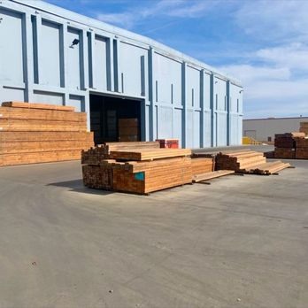 Builders FirstSource - An outdoor lumber yard featuring stacks of bundled wooden planks organized in piles. The area is adjacent to a large industri... - Building Supplies Near Me - Woodland, California