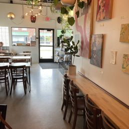 The image shows the interior of a coffee shop with a minimalist design. Hanging plants are suspended from the ceiling, and artwork is displayed on the wall. There are multiple seating arrangements, including a bar-height counter with chairs along the right side and tables with chairs on the left. The entrance door is visible at the back, with windows allowing natural light to enter. A person is seated at a table near the window, and the space has a modern, cozy atmosphere.