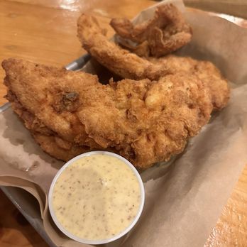 Thirsty Beaver - Chicken tender plate - A tray contains several pieces of fried chicken tenders with a golden-brown, crispy coating. A small cup of mustard-based dipping sauce with visible mustard seeds is placed in front of the chicken. The tray is lined with parchment paper and rests on a wooden table, with a background of wooden chairs and tables in a casual dining setting. - Burgers Near Me - Smithfield, Rhode Island