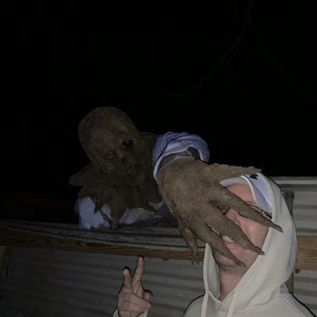 Terror Maze - A person wearing a white hoodie is taking a selfie with a creature that has a mask resembling a zombie or monster, featuring a decayed face and long fingers. The creature appears to be leaning over a wooden railing, and the background is dark, indicating it is nighttime. The person is making a peace sign with their hand. - Haunted Houses Near Me - Springfield, Ohio