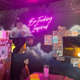 a neon sign above a wooden table - A neon sign with pink cursive text saying 'Be Fucking Inspired' is mounted on a dark wall decorated with artwork and brick accents.